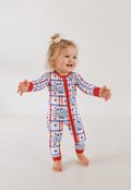 Smiling toddler standing in the In My Baby Era long sleeve bamboo zippy, a white romper with red and blue plaid, stars and "In my baby era" lettering, with red ribbed trim at the neck, cuffs and ankles.