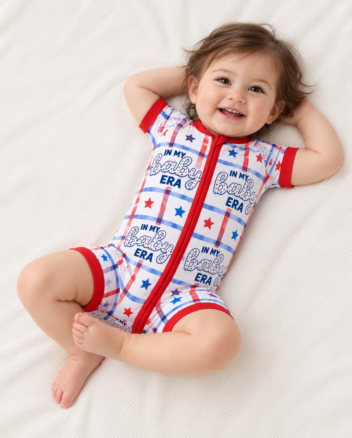 Smiling baby lying on a white blanket in the In My Baby Era short sleeve bamboo zippy, a red, white and blue plaid romper with stars, baby script and red trim.