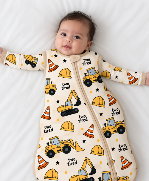 Baby lying on a white sheet wearing the Two-Tired Construction long-sleeve bamboo sleep sack with yellow excavators, dump trucks and orange traffic cones on a cream print