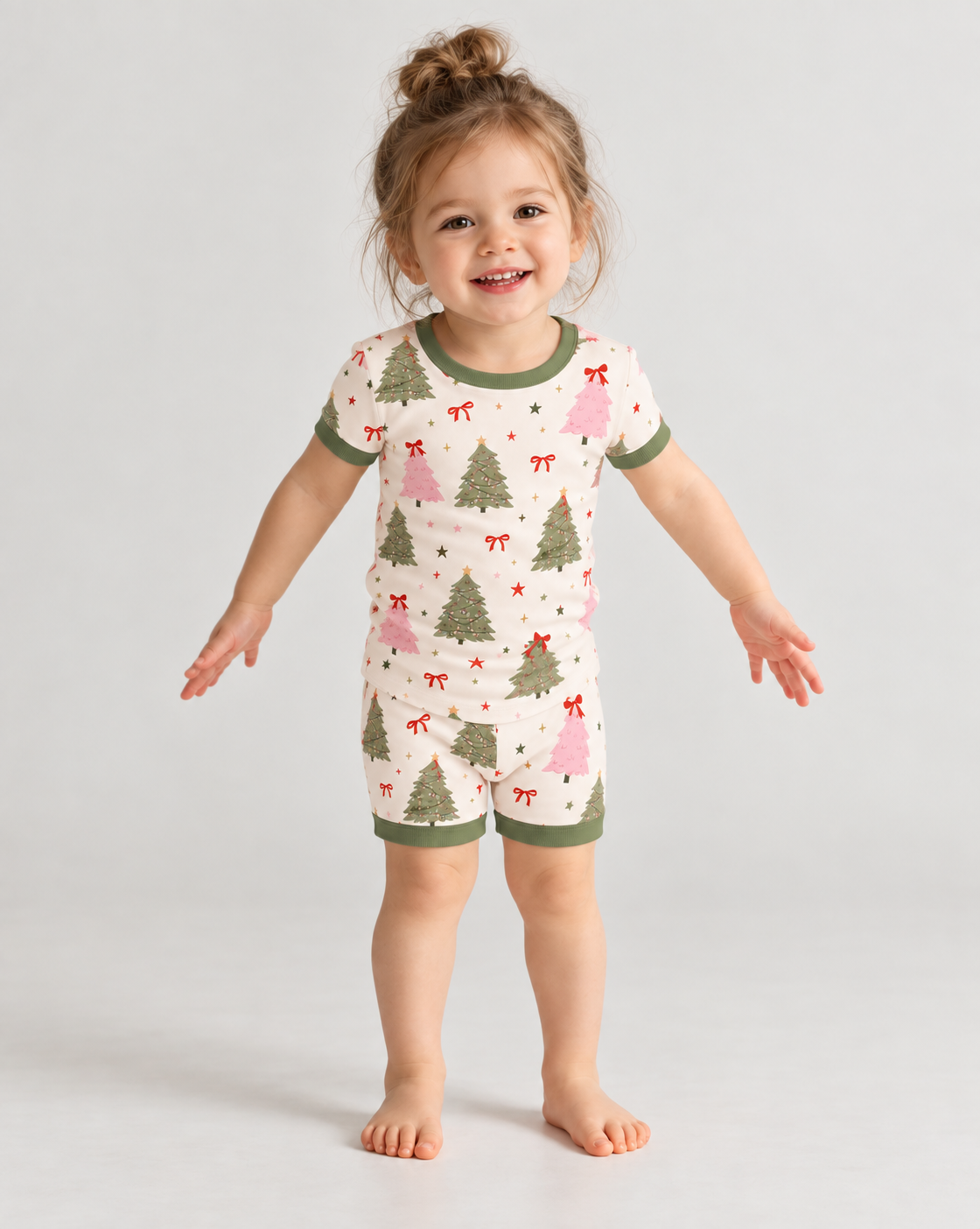 Smiling toddler in Christmas Tree short sleeve bamboo pajamas, a cream tee and shorts with sage green and pink trees, red bows and sage green trim