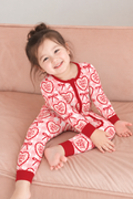 Smiling toddler on a pink sofa wearing the Jesus Loves You bamboo zippy with cream hearts and red bows on pink gingham and red cuffs