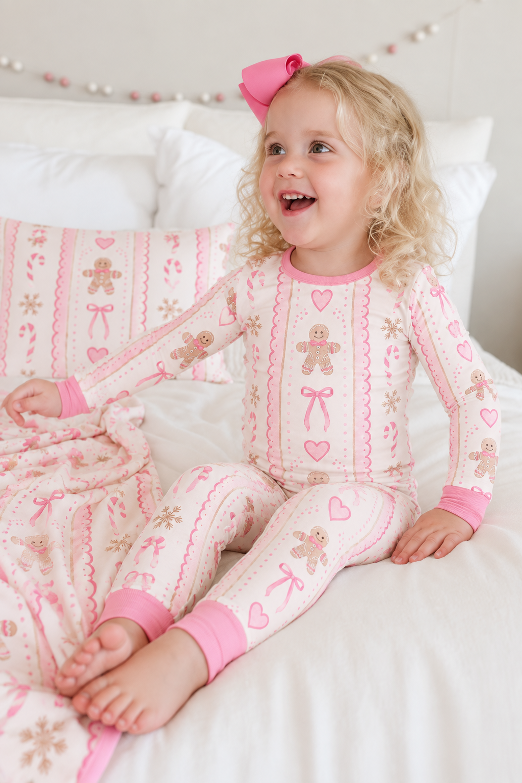 Smiling toddler girl with a pink hair bow sitting on a bed in the Pink Gingerbread long sleeve bamboo pajama set with pink cuffs and ankle bands