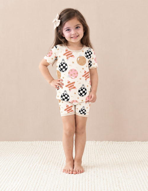 Girl wearing Merry & Bright short sleeve bamboo pajamas with checkerboard ornaments, blush and gold baubles and pink-striped stars on cream, with beige trim.