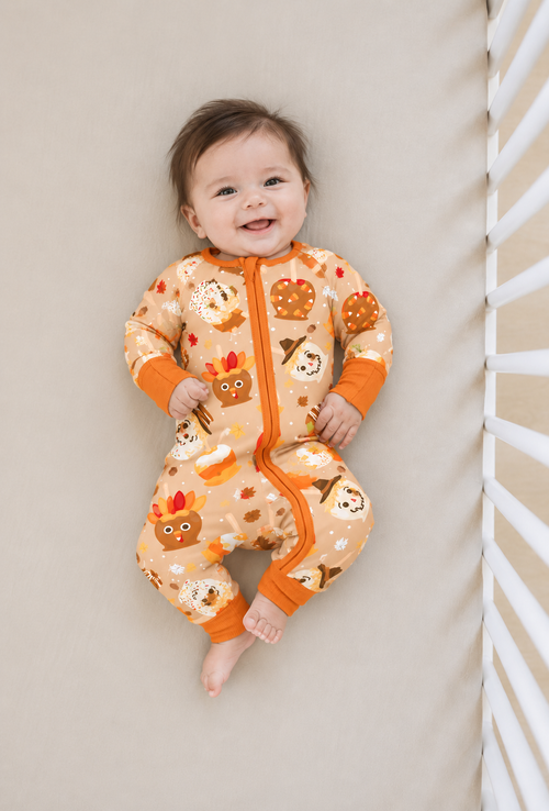 Smiling baby lying in a crib wearing the Gobble Apples bamboo zippy, a camel print of turkey and scarecrow caramel apples with a burnt orange zip and cuffs