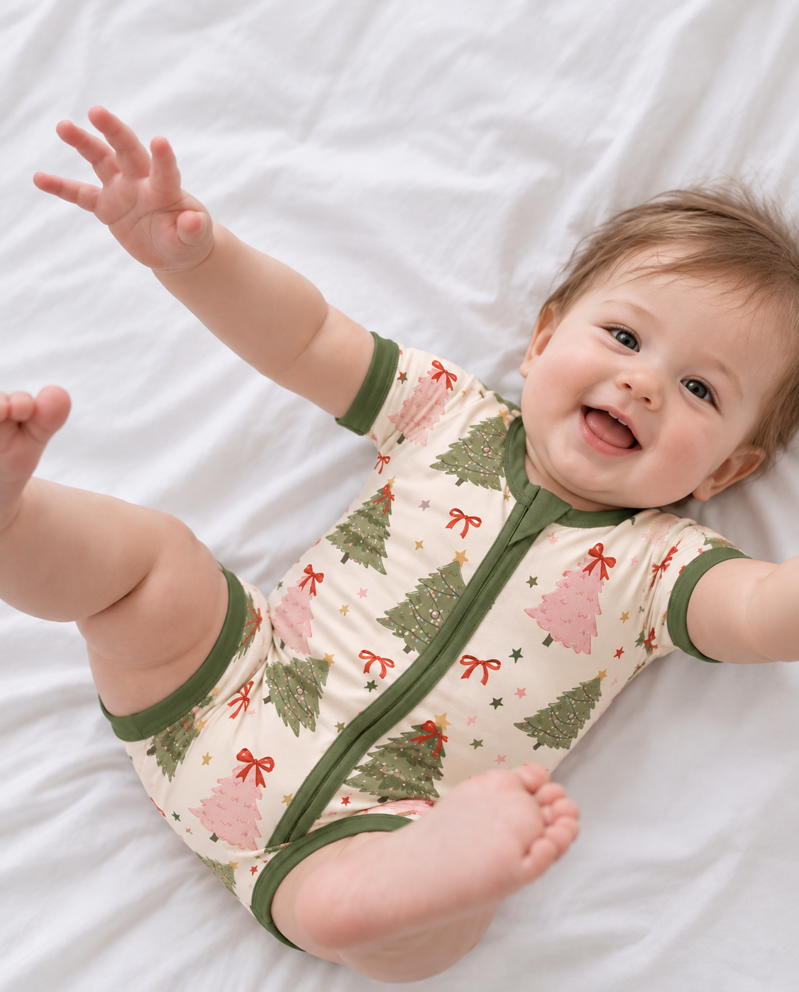 Smiling baby lying on a white bed in the Christmas Tree short sleeve bamboo zippy, with pink and sage green trees and red bows on cream