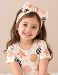 Smiling toddler girl wearing the Merry & Bright bamboo bow headband with checkerboard ornaments and blush and gold stars, paired with the matching outfit