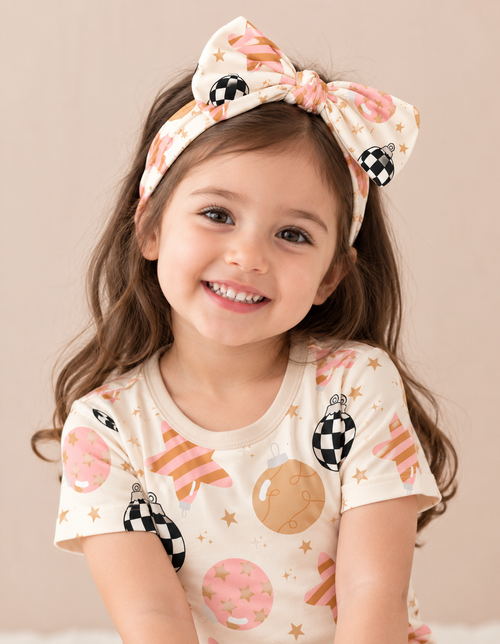 Smiling toddler girl wearing the Merry & Bright bamboo bow headband with checkerboard ornaments and blush and gold stars, paired with the matching outfit