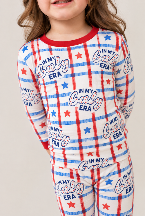 Girl wearing the In My Baby Era long sleeve bamboo pajamas, a white set with red and blue plaid, stars and "In my baby era" lettering, with a red ribbed crew neck and cuffs.