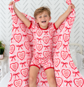 Laughing boy in Jesus Loves You shorts pajamas, cream hearts and red bows on pink gingham with red ribbed trim, holding up a blanket in the same print