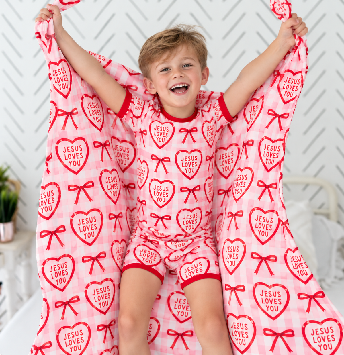 Laughing boy in Jesus Loves You shorts pajamas, cream hearts and red bows on pink gingham with red ribbed trim, holding up a blanket in the same print