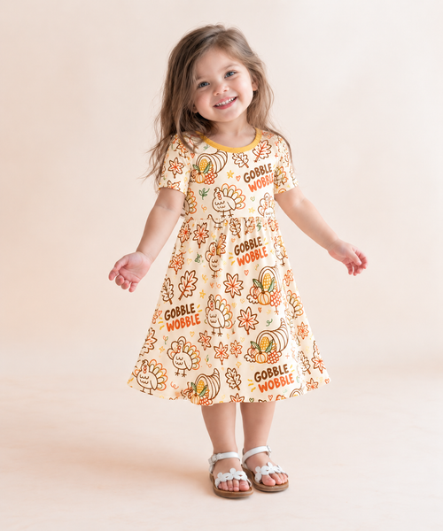 Toddler girl in the Gobble Wobble short sleeve bamboo dress with doodled turkeys and orange and brown Gobble Wobble lettering on cream