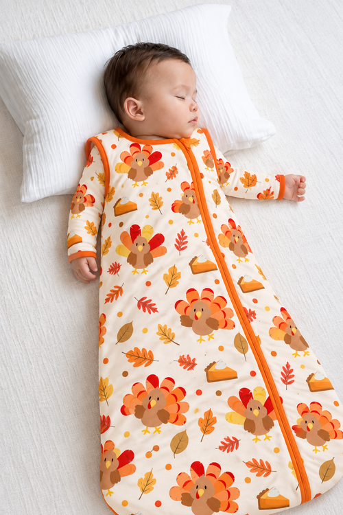Baby sleeping on a white pillow in the Turkey & Pie bamboo sleep sack, cream with turkeys, pumpkin pie slices and autumn leaves, orange zipper and trim