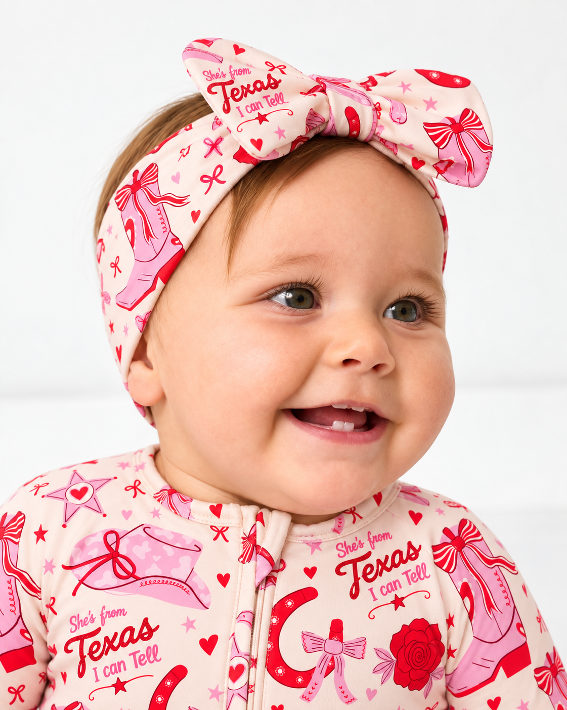 Smiling baby in the Little Texan bamboo bow headband and matching zippy, pink cowgirl boots and red roses print on cream