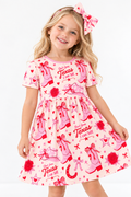 Smiling girl in a long sleeve Little Texan bamboo twirl dress and matching hair bow, pink cowgirl boots and red roses print