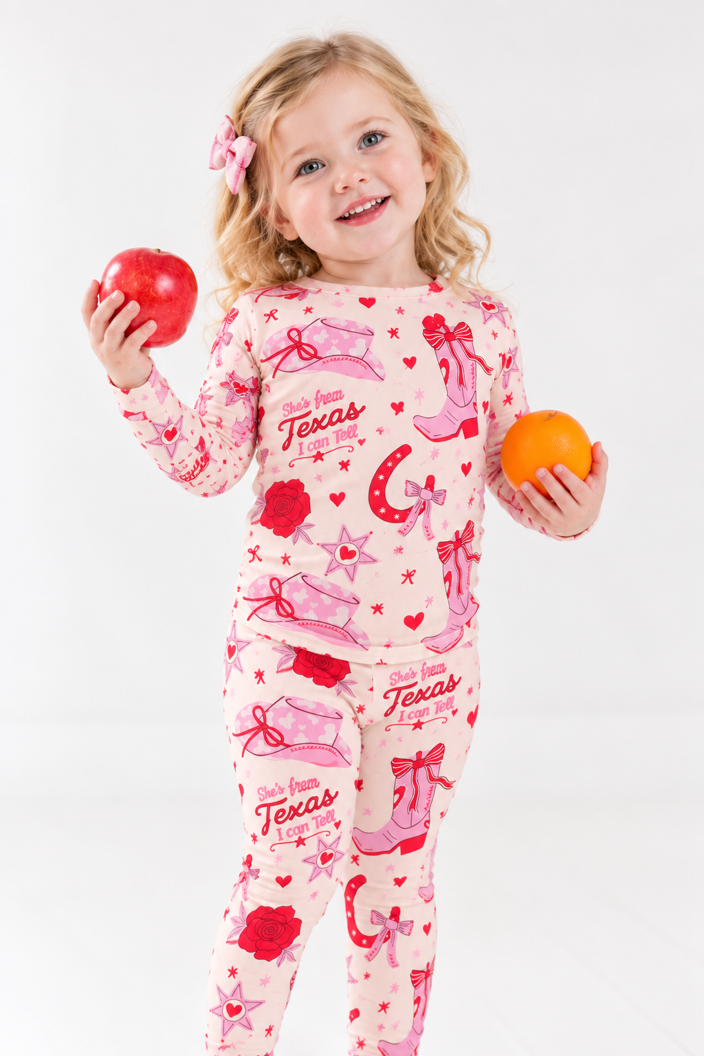 Smiling toddler girl in the Little Texan long sleeve bamboo pajama set, with pink cowgirl boots, cowboy hats and red roses on cream