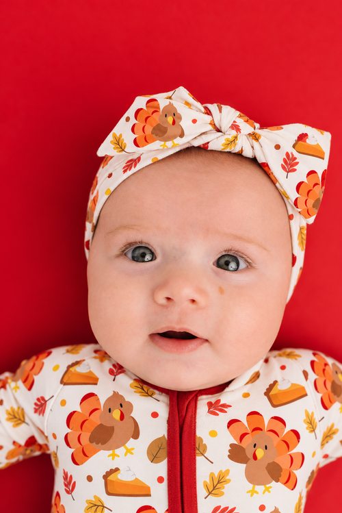 Toddler girl wearing the Turkey & Pie bamboo bow headband with a big bow, cream with turkeys and autumn leaves, paired with the matching zippy with orange zipper