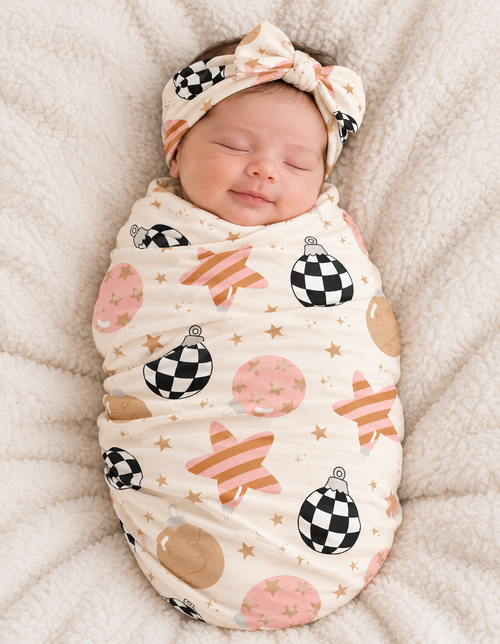 Sleeping newborn wrapped in the Merry & Bright bamboo swaddle blanket, with black and white checkerboard ornaments, blush and gold baubles and pink-striped stars on cream.