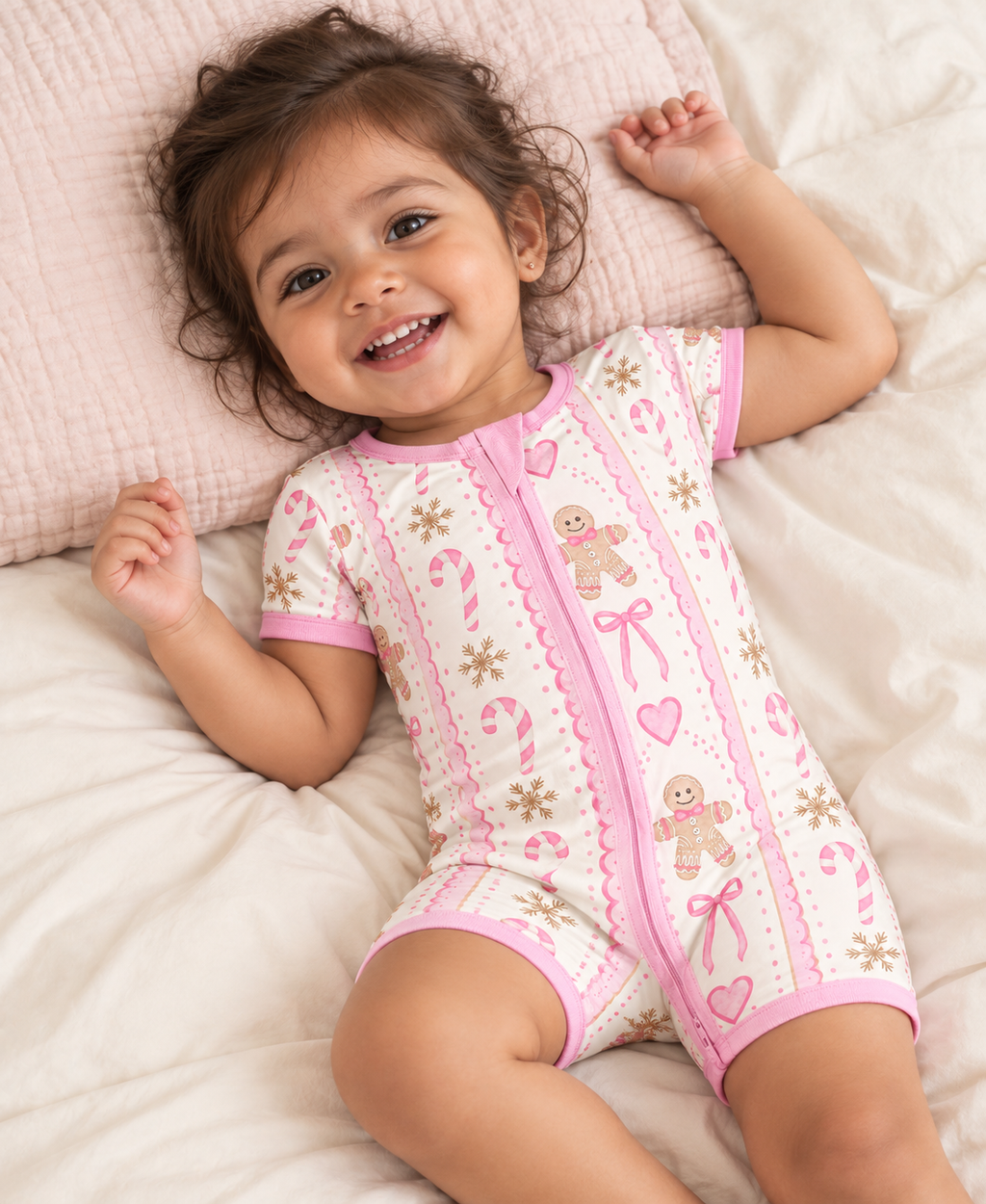 Smiling baby lying on a bed in the Pink Gingerbread Bamboo Short Zippy with gingerbread men, candy canes and pink trim.