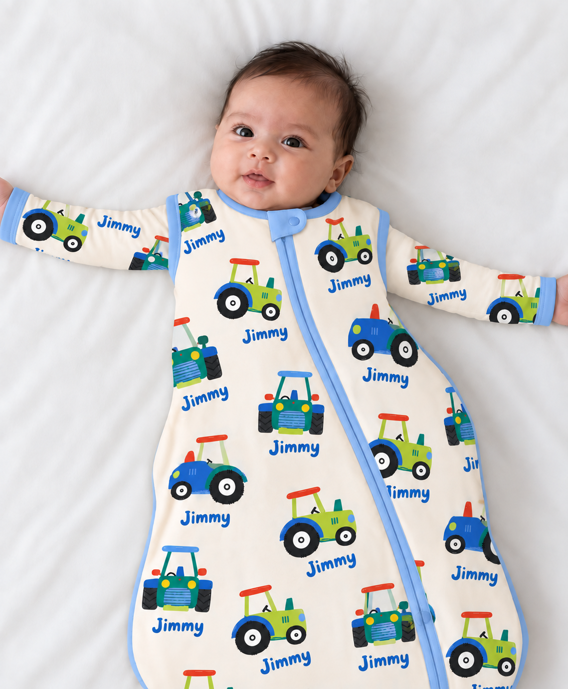 Baby in a cream bamboo sleep sack printed with blue, green and red tractors and the name Jimmy, with royal blue trim and front zip