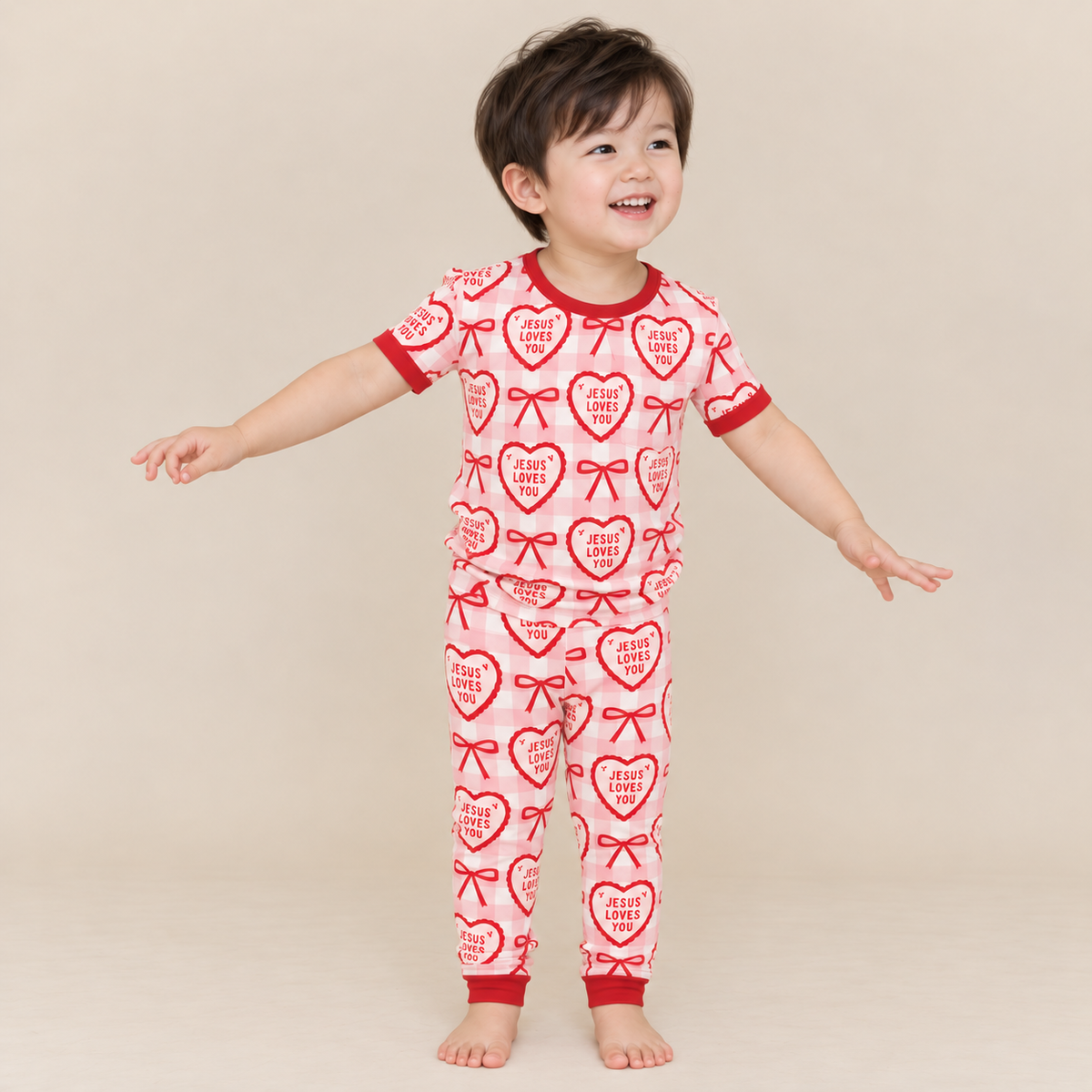 Smiling toddler in Jesus Loves You short sleeve pajamas with long pants, cream hearts and red bows on pink gingham, red ribbed neckline, sleeve hems and ankle cuffs