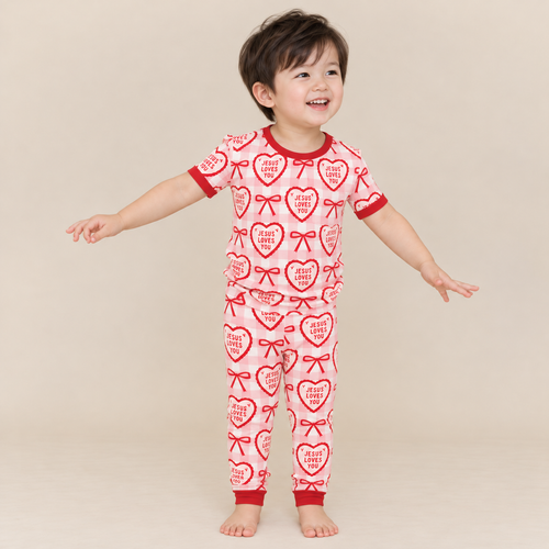 Smiling toddler in Jesus Loves You short sleeve pajamas with long pants, cream hearts and red bows on pink gingham, red ribbed neckline, sleeve hems and ankle cuffs